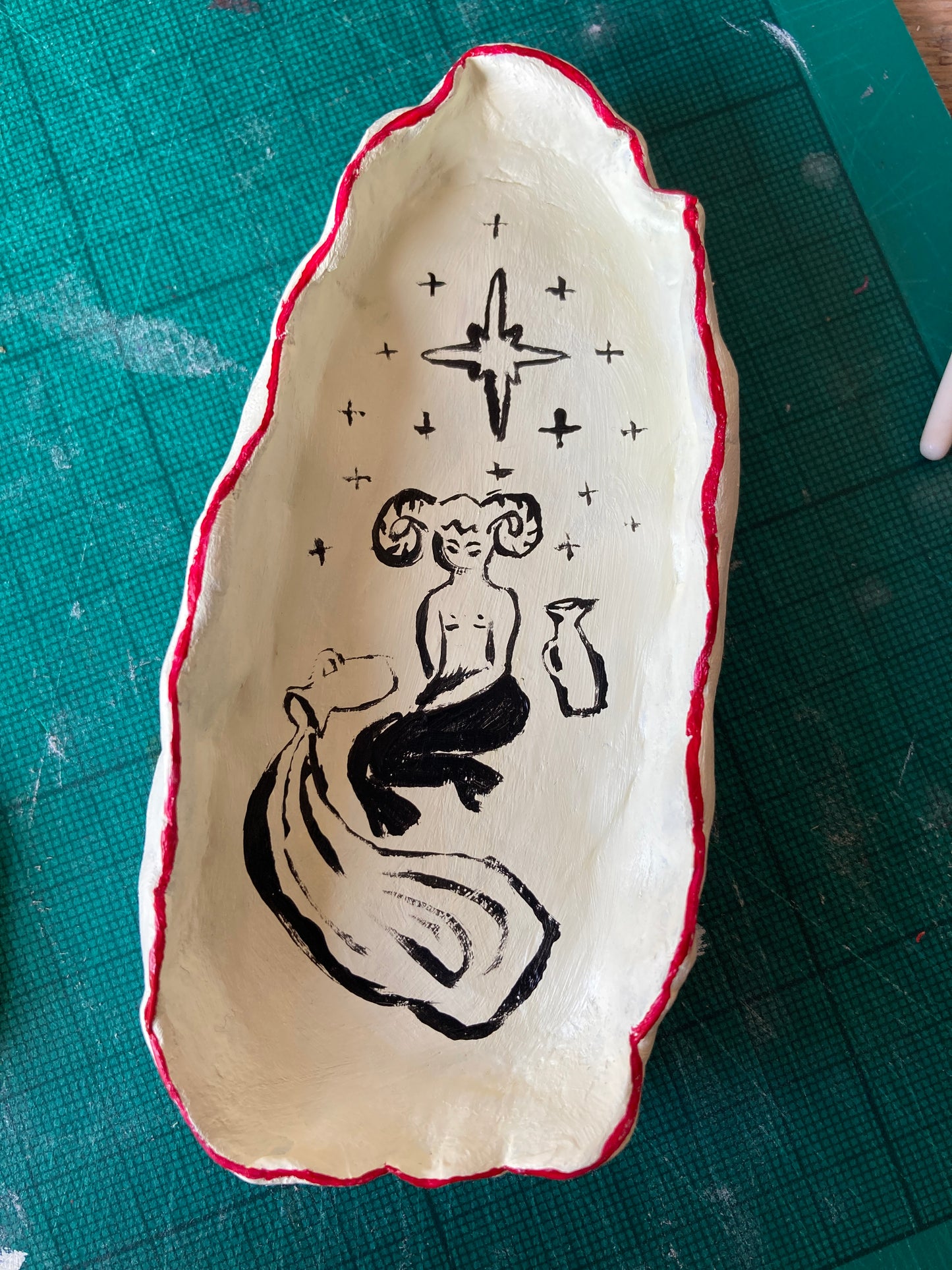 A small ceramic bowl with red rim against an off white background with a satyr, vase, spilt vase and stars on a green cutting matt.