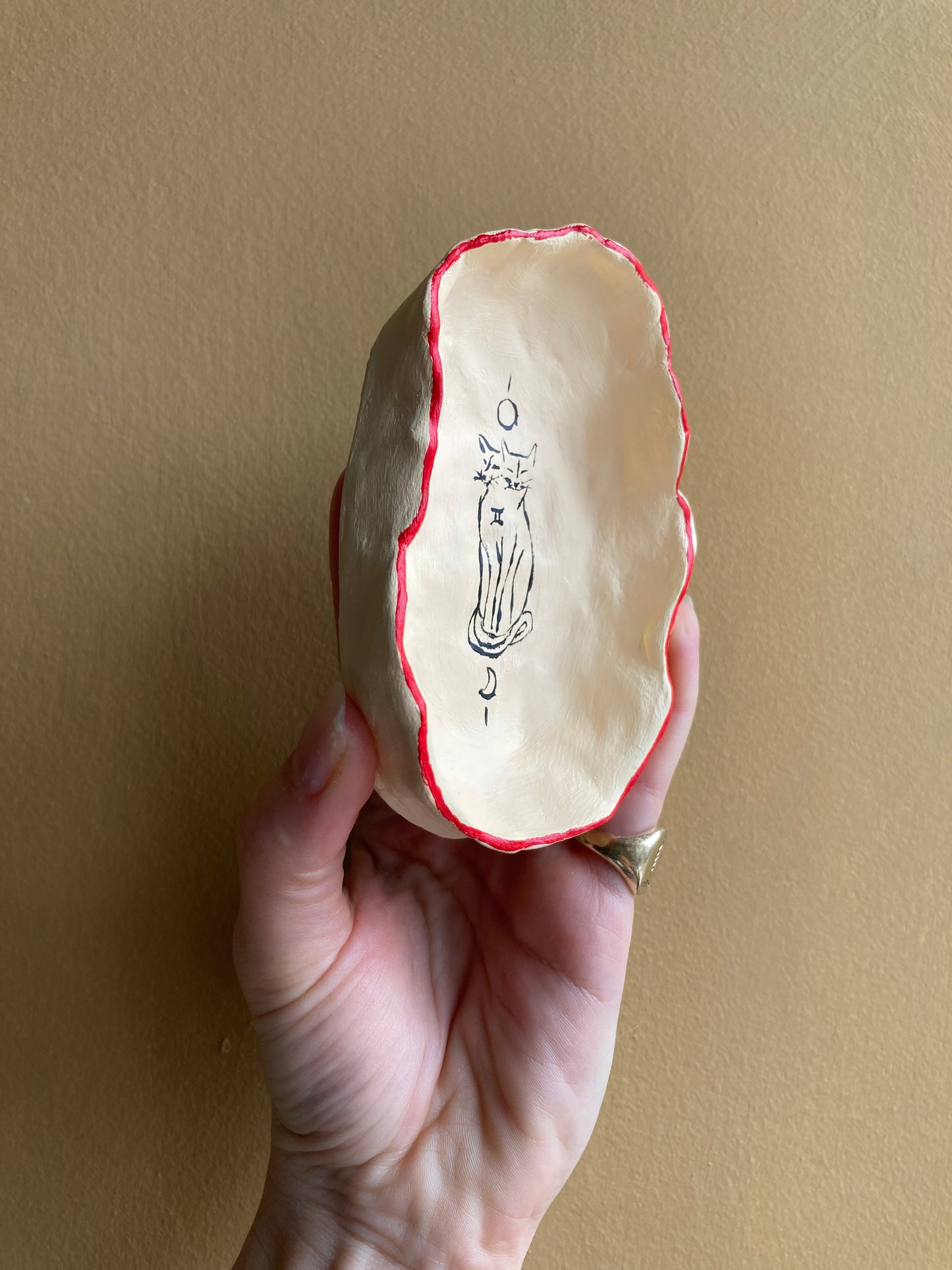Hand holding a small ceramic bowl with red rim against an off white background with a gemini two-headed cat and a sun and moon.