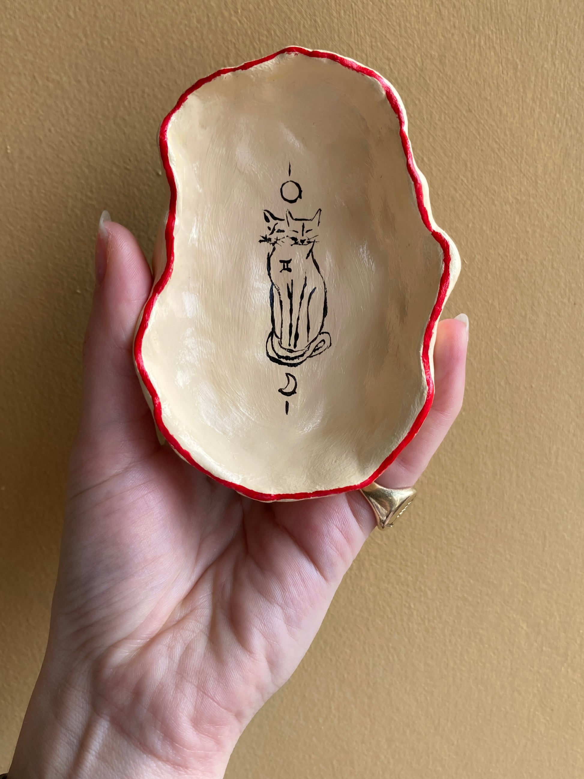 Hand holding a small ceramic bowl with red rim against an off white background with a gemini two-headed cat and a sun and moon.